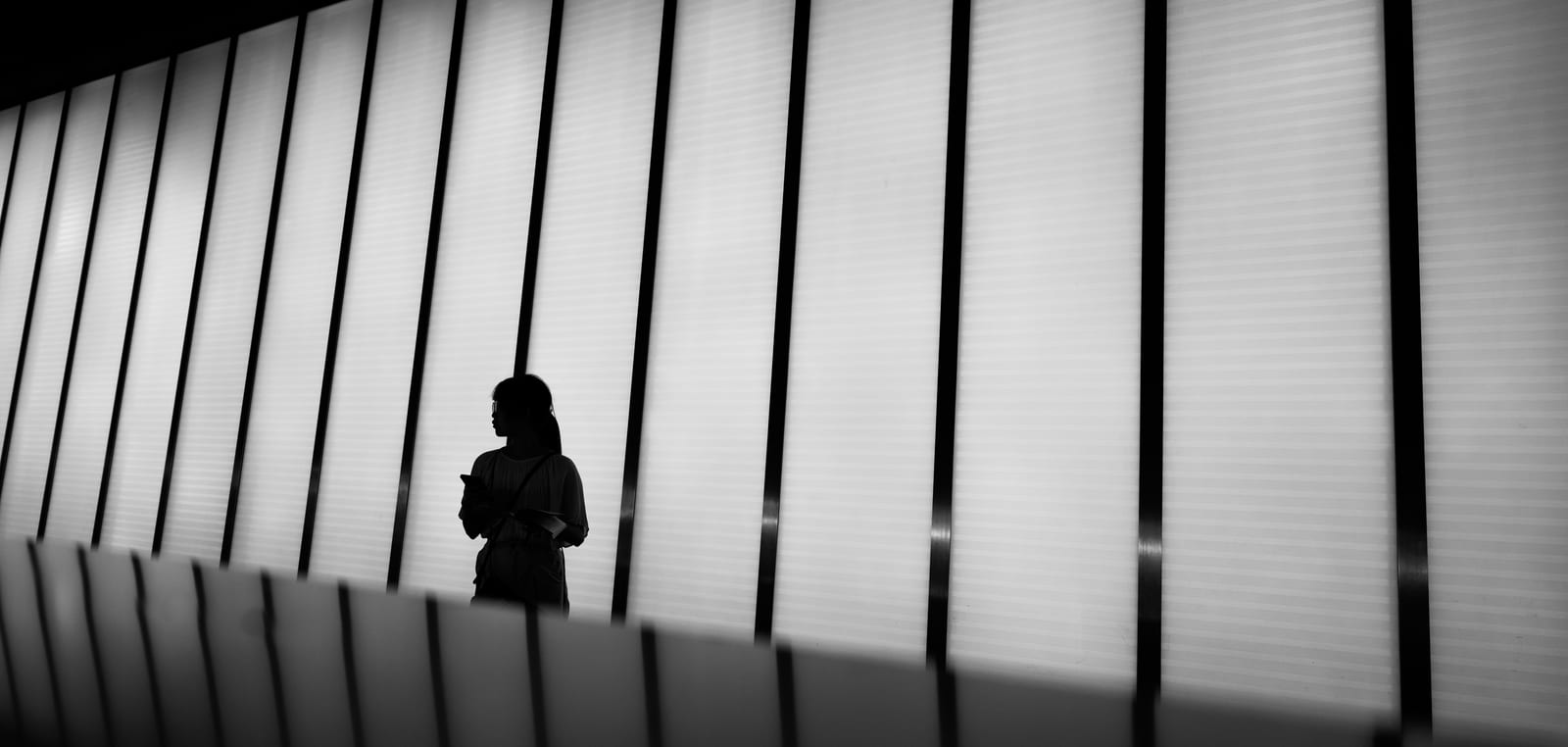 Black-and-white photograph of a woman in silhouette holding a phone, standing against a tall backlit louvered wall that fills the frame with vertical bands of light.
