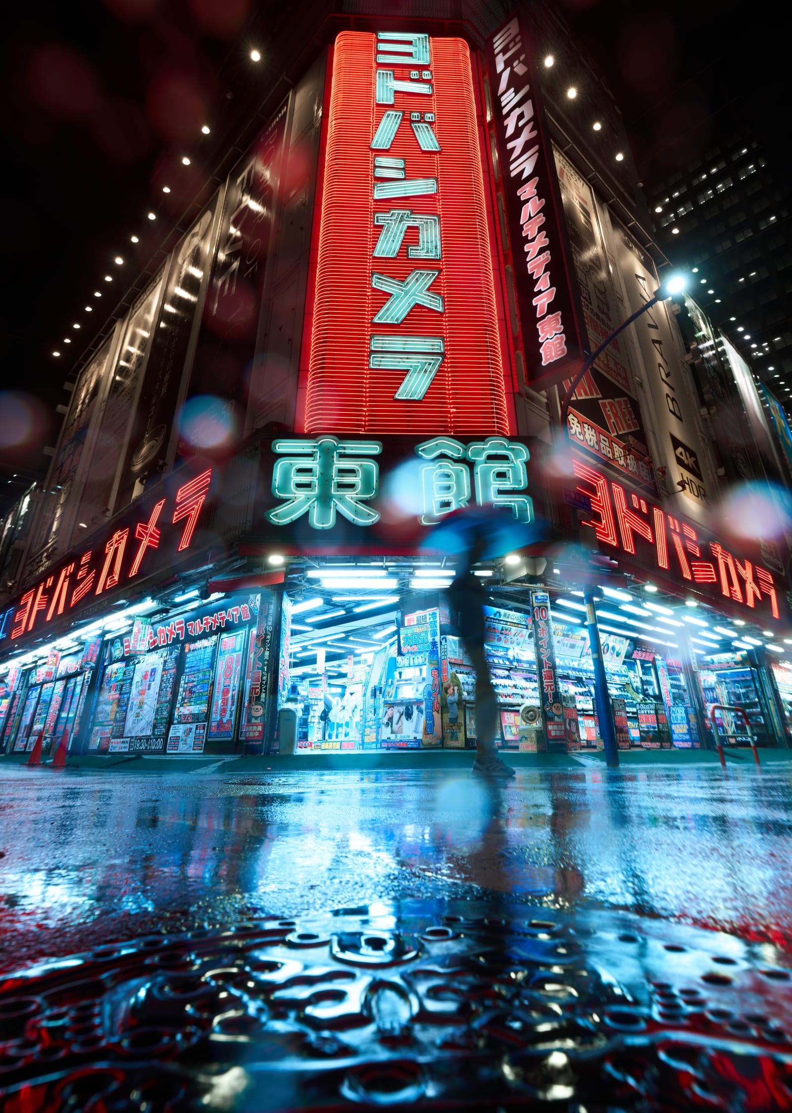 Low-angle night view of the Yodobashi Camera Multimedia East building in Shinjuku, its red and cyan neon signage reflected in a rain-soaked street, a blurred figure under an umbrella crossing the foreground.